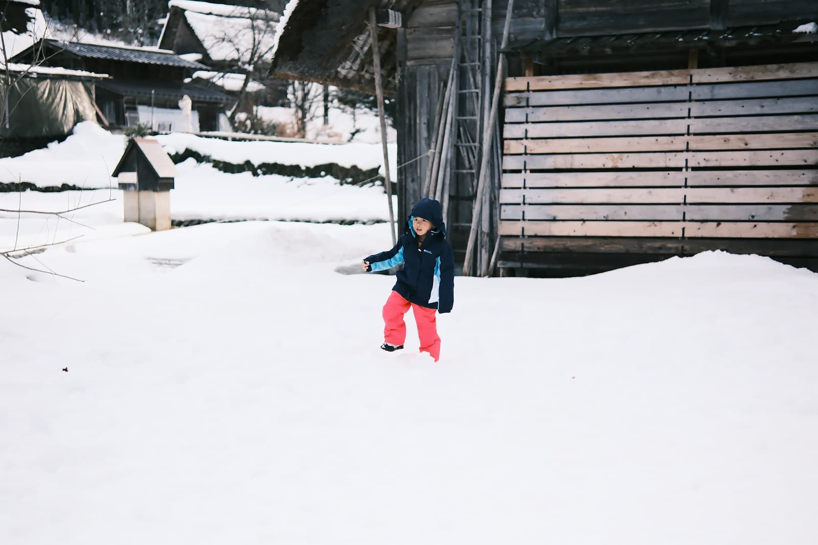 Un enfant joue dans la neige, près de bâtiments en bois.