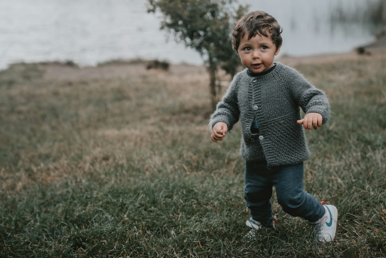 Un garçon en pull gris et jean bleu, debout dans l'herbe.